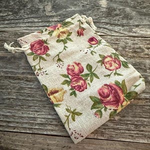 May include: A rectangular drawstring pouch made of a light beige, textured fabric featuring a repeating pattern of pink and yellow roses with green leaves and stems. The bag is closed at the top with white twisted cord drawstrings on both sides.