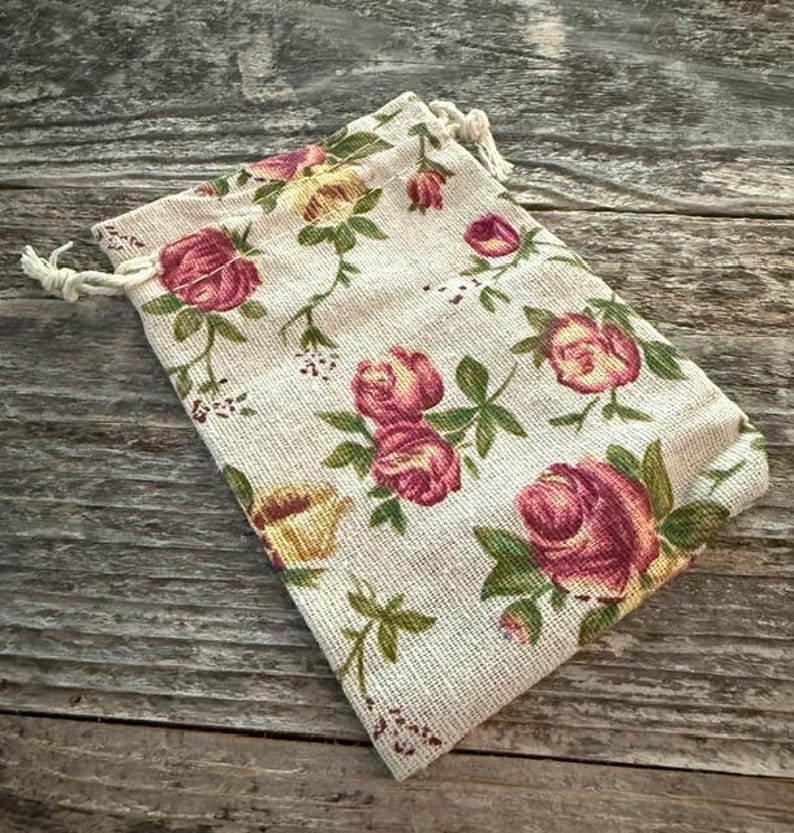 May include: A rectangular drawstring pouch made of a light beige, textured fabric featuring a repeating pattern of pink and yellow roses with green leaves and stems. The bag is closed at the top with white twisted cord drawstrings on both sides.