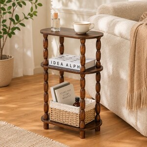May include: A three-tier oval side table made of dark brown wood with turned spindle legs. The top shelf holds a glass perfume bottle and a white ceramic mug. The middle shelf displays a book titled IDEA ALPH. The bottom shelf holds a woven basket.