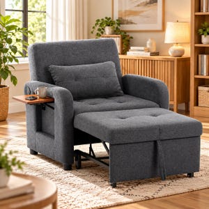 May include: A charcoal gray convertible chair bed featuring a tufted backrest, a matching rectangular throw pillow, and a pull-out ottoman base. A small wooden side table is attached to the left armrest, holding a ceramic mug and a phone.