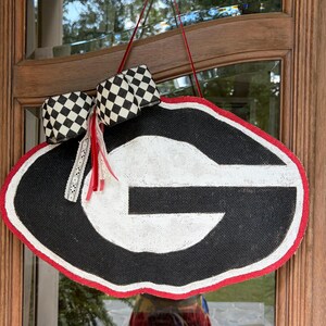 May include: A black and white oval-shaped door hanger featuring a large white letter G on a black background with a red border. The piece is accented with a black and white checkered ribbon bow and a thin red ribbon for hanging on a door.