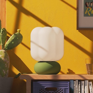 May include: A table lamp featuring a rounded, matte olive green base and a white, translucent, pumpkin-shaped glass shade. The lamp sits on a wooden surface next to a potted cactus and a clear glass of water, with a framed print visible nearby.