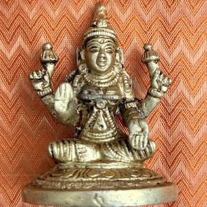 Peut inclure: Une figurine en métal doré représentant une divinité à quatre bras assise en tailleur sur une base circulaire. La figure porte une coiffe et des bijoux ornés, avec deux mains supérieures tenant de petits objets et deux mains inférieures ouvertes.
