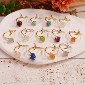 May include: A collection of gold-toned rings featuring a crescent moon design on one end and a raw, uncut gemstone on the other. The stones display various colors including deep red, turquoise, light blue, green, purple, orange, pink, and yellow.