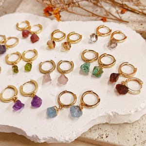 May include: A collection of gold-toned hoop earrings, each featuring a single raw, uncut gemstone pendant. The stones display various colours including purple, green, pink, blue, clear, and brown. The earrings are arranged in pairs on a white surface.