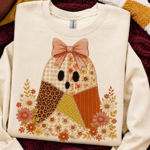 May include: A cream-colored crewneck sweatshirt featuring a central graphic of a ghost figure composed of various patterned patches including gingham, leopard print, and floral designs, topped with a large bow and surrounded by colorful flowers.