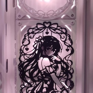 Könnte beinhalten: Eine weiße Lüfterabdeckung für Computer mit einem dekorativen Schleifendesign, positioniert über einem schwarzen, lasergeschnittenen Lüftergitter, das eine Anime-Figur zeigt, die von komplizierten Herz- und Bandmustern umrahmt wird.