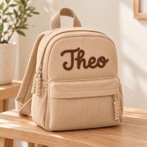 Può includere: Uno zaino in velluto a coste beige con il nome Theo ricamato in corsivo marrone scuro sulla tasca anteriore. La borsa include un manico superiore, spallacci regolabili e tiralampo intrecciati in tinta con il tessuto dello zaino.