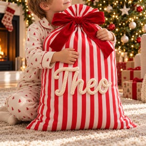 May include: A large fabric gift sack featuring vertical red and white stripes, cinched at the top with a large red velvet bow. The name Theo is written across the front in thick, cream-colored knitted yarn lettering.