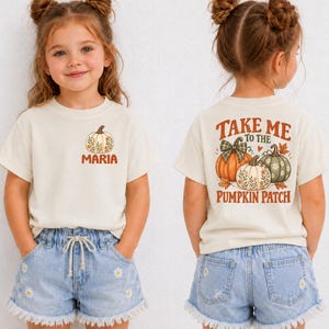 May include: A cream-colored short-sleeve t-shirt featuring a small pumpkin graphic and the name MARIA on the front left chest. The back displays a large graphic of three pumpkins with the text TAKE ME TO THE PUMPKIN PATCH in orange letters.