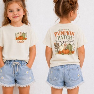 May include: A cream-colored t-shirt featuring a small pumpkin graphic with the name LAURA on the front and a large pumpkin patch graphic with the text PUMPKIN PATCH CREW on the back, paired with light blue denim shorts with white daisy embroidery.