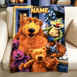 Pode incluir: Uma manta retangular de lã com um urso grande castanho, um urso laranja mais pequeno, duas criaturas roxas, uma criatura verde e uma pequena criatura azul. A palavra NAME está impressa a amarelo no topo sobre um fundo de estante azul.