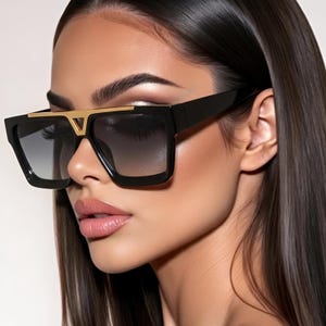 May include: Oversized square sunglasses featuring a thick black frame and gradient gray lenses. The bridge is accented with a prominent gold-toned metal V-shaped detail that connects the upper frame, providing a bold and structured appearance.
