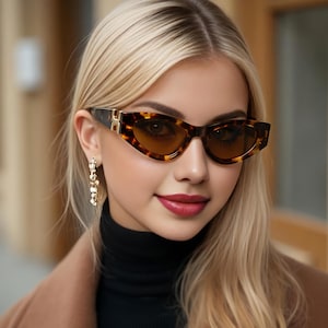 May include: Cat-eye sunglasses featuring a brown tortoiseshell pattern frame with dark tinted lenses and a gold-toned metal accent on the temple hinges. The eyewear has a pointed outer edge design and a smooth, polished finish on the plastic frame.