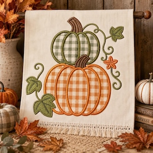 May include: A cream-colored fabric wall hanging featuring an embroidered design of two stacked pumpkins. The top pumpkin has a green and white gingham pattern, while the bottom pumpkin has an orange and white gingham pattern. Green vines and leaves surround the pumpkins, and the bottom edge is finished with a fringe trim.