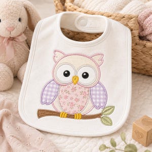 May include: A white baby bib featuring an embroidered owl design. The owl has a pink floral patterned belly, purple gingham wings, and large black eyes. It is perched on a brown branch with green leaves. The bib has a snap closure at the neck.