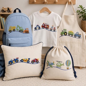 Puede incluir: Una mochila azul claro, una camiseta blanca, un delantal crema, un cojín crema y una bolsa con cordón crema. Cada artículo presenta vehículos coloridos bordados, incluyendo camiones de construcción, vehículos de emergencia, tractores y aviones.