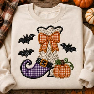 May include: A cream-colored crewneck sweatshirt featuring an embroidered design of a witch boot with a purple and white checkered toe, a floral patterned shaft, and an orange and white checkered bow. Three black bats and an orange pumpkin are included.