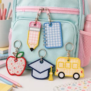 May include: Five embroidered keychains featuring school-themed shapes: a pink gingham pencil, a blue gingham notebook, a floral apple, a blue striped graduation cap, and a yellow gingham school bus, all attached to silver metal key rings.