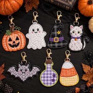 May include: A collection of seven embroidered Halloween-themed keychains featuring a jack-o'-lantern, a ghost, a witch hat, a cat, a bat, a potion bottle, and candy corn. Each keychain has a gold-tone metal lobster clasp attachment.