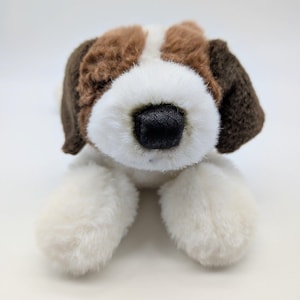 May include: A plush dog toy featuring soft white, brown, and dark brown fur. The stuffed animal has a prominent white muzzle, a black nose, and floppy dark brown ears. The design shows the dog in a lying position with its front paws extended forward.