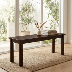 May include: A rectangular dark brown wooden bench with four straight legs. On the flat surface of the bench sits a small cream-colored ceramic vase containing dried beige grasses and a stack of three light-colored books with neutral covers.