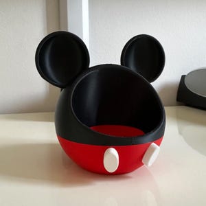 May include: A black and red spherical container featuring two large circular ears on top, resembling a cartoon character head. The lower portion is red with two white oval accents on the front, while the upper portion is black with a wide opening.