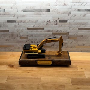 May include: A yellow and black CAT excavator scale model mounted on a rectangular wooden base. The base features a gold-colored metal plaque with engraved text that reads "Midstream Stanley" and "Certified Professional Level".
