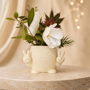 May include: A cream-colored ceramic planter shaped like a character with a smiling face and two arms making peace signs. The planter holds a bouquet of white flowers, green leaves, and dark foliage, creating a cheerful and decorative display.