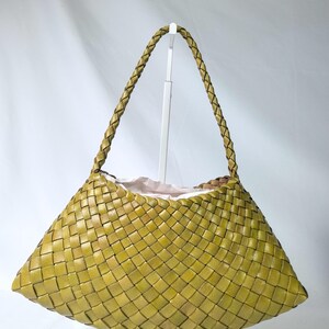 Puede incluir: Un bolso de mano color verde chartreuse con una construcción de cuero tejido en un patrón de cestería. El bolso tiene una base ancha y curva, y una sola correa de hombro de cuero trenzado sujeta a las esquinas superiores del cuerpo.