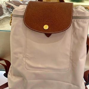 May include: A beige nylon backpack featuring a brown leather flap with a gold-tone snap button closure. The backpack includes a top zip, adjustable brown straps, and a rectangular front pocket design with visible stitching on the main body.