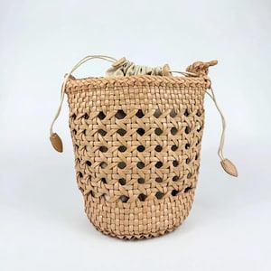 May include: A light tan bucket bag featuring an intricate woven lattice exterior pattern. The bag includes a drawstring closure with thin leather cords and a light beige fabric interior lining visible through the open weave of the structure.