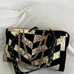 Può includere: Una borsa rettangolare caratterizzata da un motivo a chevron che alterna pannelli in tessuto nero e pannelli con texture effetto pelle di serpente. La borsa include una tracolla a catena in metallo dorato e un ampio cinturino in pelle nera con occhielli in metallo argentato.