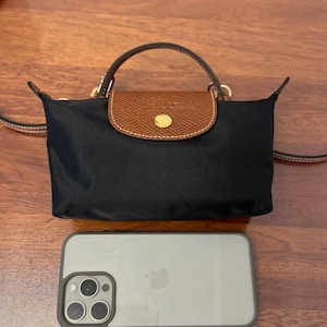 May include: A black nylon handbag with a brown leather flap featuring a gold-tone snap button closure and a top handle. The bag includes a thin shoulder strap and is positioned above a smartphone to provide a visual scale of its compact size.