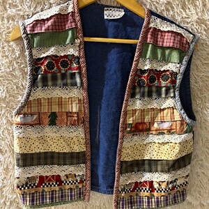 May include: A sleeveless vest featuring a patchwork design of horizontal fabric strips in various patterns including sunflowers, pumpkins, and barns, accented with white lace trim. The vest has a solid dark blue back and a brown checkered trim.