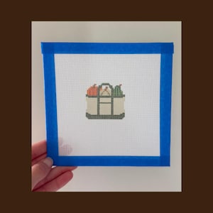 Könnte beinhalten: Eine quadratische Stickleinwand mit einem kleinen gestickten Motiv einer beigen Tragetasche, gefüllt mit einem orangefarbenen Kürbis und einer grünen Birne. Die Leinwand ist an allen vier Seiten mit blauem Malerkrepp umrandet, Text Lee Stitchery.