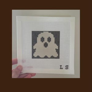 May include: A square needlepoint canvas featuring a pixelated ghost design in light beige against a solid black background. The canvas is framed with white tape on all four sides. The letters L S are stitched in black in the bottom right corner.