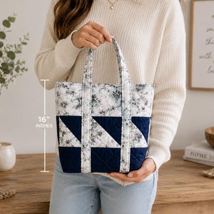 以下が含まれることがあります： ネイビーブルーと白の花柄に幾何学的な三角形のデザインが施されたキルティングトートバッグ。2本の布製ハンドルが付いており、前面に表示されたテキストと測定線により高さが16インチであることが示されています。