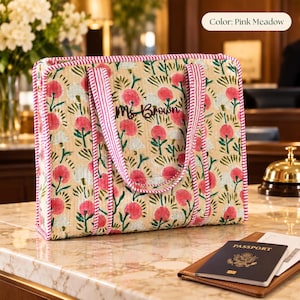 Op de afbeelding: Een rechthoekige gewatteerde tas van beige stof met een herhalend bloemenpatroon in roze en groen. De tas heeft roze en wit gestreepte hengsels en biezen, met de naam Ms. Brown in donker handschrift geborduurd op de voorzijde.