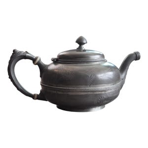 May include: A dark gray metal teapot with a rounded body, a curved handle, and a spout. The surface features engraved floral patterns and a bird motif, with a horizontal rope-like texture band encircling the center and a lid with a finial.