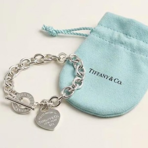 May include: A silver chain link bracelet with a toggle clasp and a heart-shaped charm, both engraved with Tiffany & Co. branding. The bracelet is accompanied by a light blue drawstring pouch featuring the Tiffany & Co. logo printed in black.