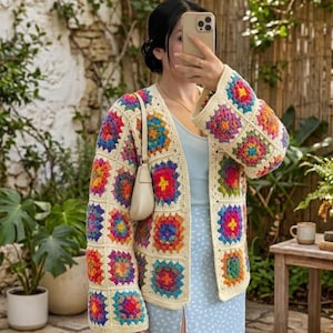 May include: A long-sleeved cardigan made of cream-colored yarn featuring a multi-colored granny square pattern in shades of red, yellow, blue, green, and purple. The garment has an open front design and a relaxed fit with wide sleeves.