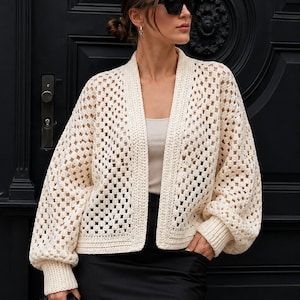 May include: An off-white crochet cardigan featuring a classic granny square pattern with an open-front design, long balloon sleeves, and ribbed cuffs. The garment is crafted with a textured, lightweight knit construction and a solid border trim.