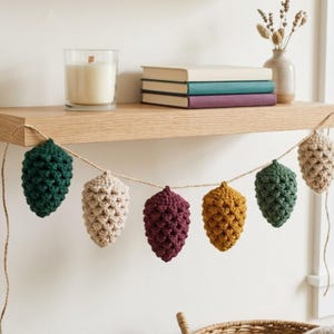 May include: A decorative garland featuring six crocheted pinecone ornaments in dark green, cream, burgundy, mustard yellow, and light beige, all suspended from a thin twine string. The ornaments have a textured, bumpy surface pattern.