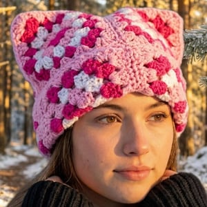 May include: A crochet beanie featuring a cat-ear silhouette, constructed from a granny square pattern in shades of light pink, dark pink, and white yarn. The hat has a textured, chunky knit appearance with pointed corners that mimic feline ears.