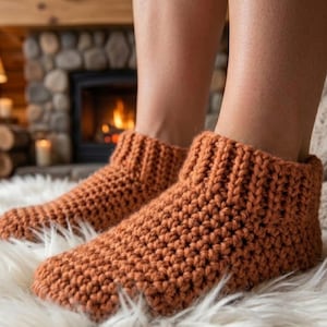 Könnte beinhalten: Ein Paar knöchelhohe Hausschuhe aus dickem, rostorangem Garn. Die Hausschuhe weisen ein strukturiertes, grobes Strickmuster mit einem gerippten Bündchen auf, das direkt über dem Knöchel sitzt und für ein gemütliches und robustes Aussehen sorgt.