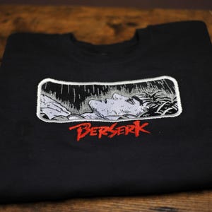 May include: A black t-shirt featuring a rectangular embroidered patch with a grayscale illustration of a person lying down in the rain. Below the patch, the word BERSERK is embroidered in a bold, jagged red font across the chest area.