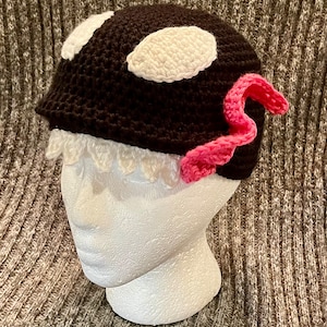 May include: A brown crocheted hat with white and pink accents. The hat has two white circles on top and a pink crocheted flower on the side.