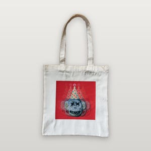 Peut inclure: Un sac fourre-tout en toile blanche avec un motif carré centré. Le motif représente un visage de singe gris texturé avec des yeux en forme de X et un sourire édenté, portant un chapeau de fête bleu clair à pois colorés sur fond rouge.