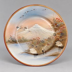 May include: A round decorative plate featuring a painted landscape of Mount Fuji, several white cranes in flight and standing in water, and colorful floral clusters. The plate has a thin red rim with a gold geometric pattern along the edge.